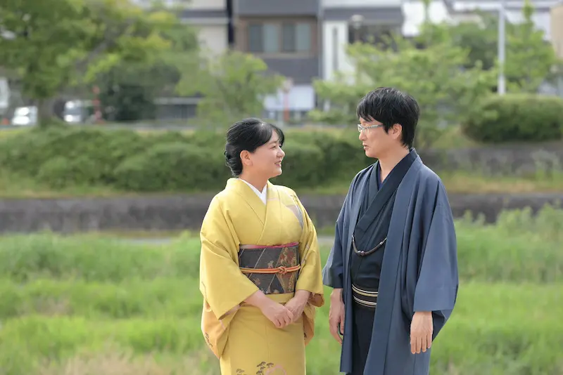 日本の伝統的な衣装「着物」を着た二人が芝生の上で微笑み合って立っている。一人は黄色の着物に柄物の帯、もう一人は青い着物に眼鏡。緑の低木とぼやけた建物が新春の情景を演出している。.