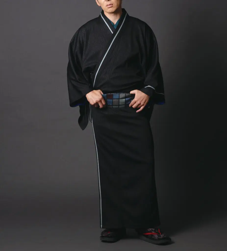 伝統的な黒の着物に柄入りの帯を締め、黒の草履を履いた人物が暗い背景の中に立っている。ブルーの縁取りと繊細な質感がアクセントとなり、新春のお祝いにぴったり。頭の一部がフレームからはみ出し、腕は両脇にある。.