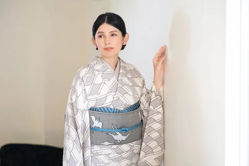 黒髪を低い位置で束ねた女性が、灰色と白の柄の着物に、白い鶴がデザインされた青と灰色の帯を締めている。彼女は室内で白い壁の近くに立ち、片手をその壁に置き、穏やかな表情で横を向いている。.