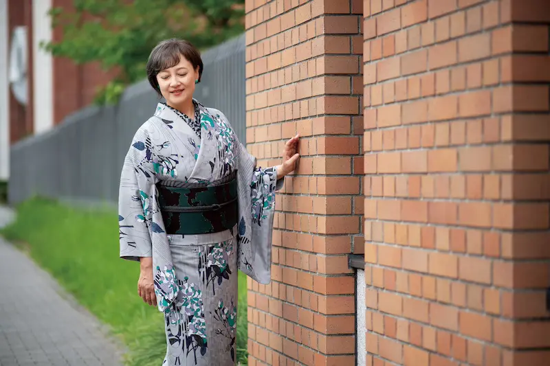 花柄のライトグレーの新春着物に濃い色の帯を締めた女性が屋外に立ち、穏やかな表情でレンガの壁にそっと触れている。彼女の横には緑の芝生と金属のフェンスが並んでいる。.