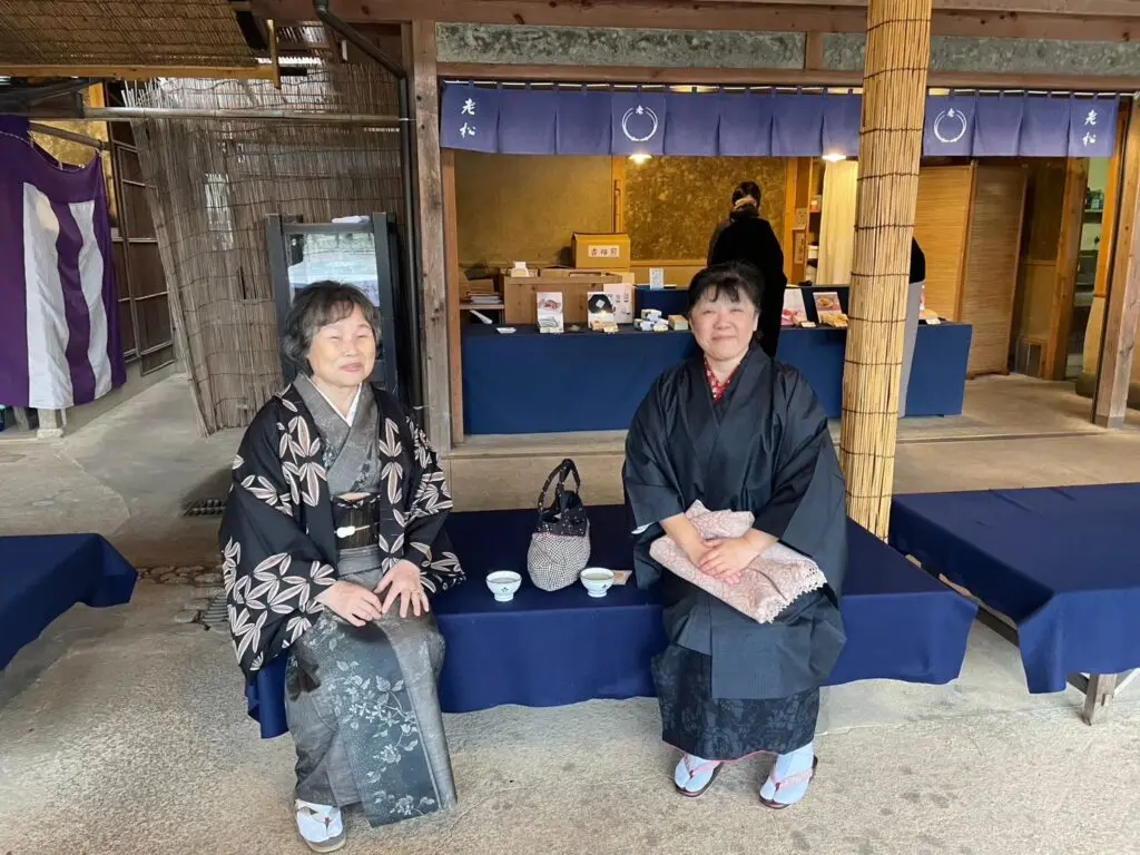 美しい着物を着た2人の女性が、青い垂れ幕と木製のファサードを持つ店の外で、青いベンチに座ってお茶を楽しんでいる。その傍らにはハンドバッグが置かれ、店内ではスタッフブログが整然と並べられた商品の中で働くスタッフの姿をとらえている。.