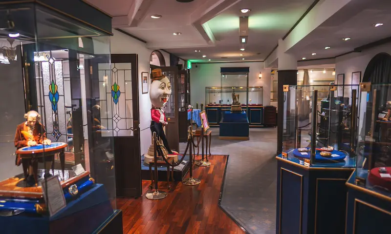 オートマタやアンティークのオブジェが展示されたガラスケースが並ぶ博物館の一室。中央には、白くて大きな人間のようなオートマタの頭部が置かれている。アーチ型の扉から自然光が差し込み、嵐山の静謐な美しさを思わせる。.