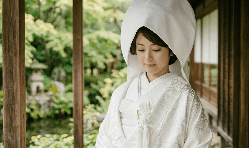 日本の伝統的な白無垢姿の女性が、縁側に立ち、下を向いて優しく微笑んでいる。背景には青々とした葉と石灯籠が描かれている。.