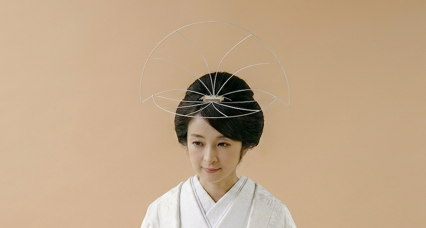 ベージュの無地を背景に立つ白無垢姿の女性は、スタイリングされた黒髪にモダンな扇形のヘッドピースをあしらっている。結婚式にふさわしい、穏やかで優しい表情は、スタッフBLOGに掲載された和の衣装の美しさを表現している。.
