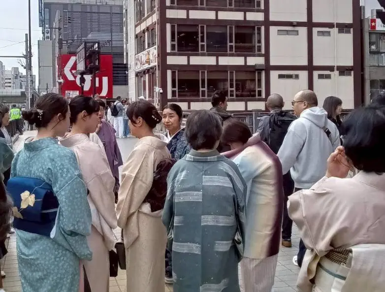 カラフルな着物を着た大勢の人々が、街の歩道で立ち話をしている。背景には近代的なビルと赤い看板。向かい合う人もいれば、カメラに背を向ける人もいる。.