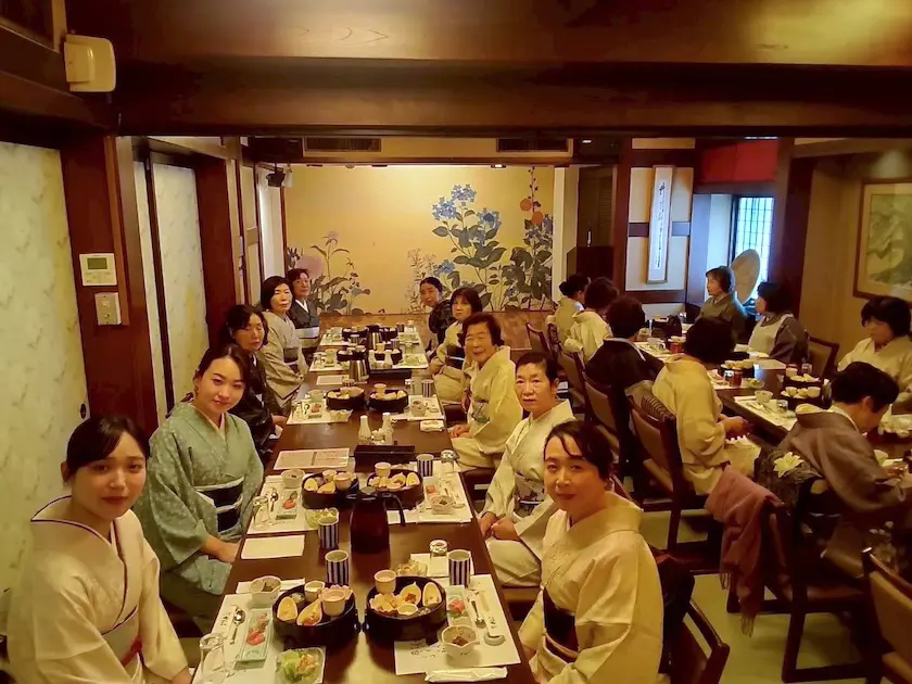 上品な着物姿の女性たちが、居心地の良い日本料理店の長テーブルに座り、一緒に食事を楽しんでいる。暖かく柔らかな照明の部屋には木の梁があり、植物が描かれた壁画があり、魅力的な雰囲気を醸し出している。.