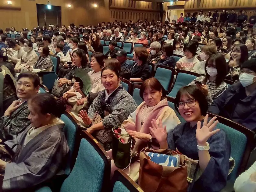 青い座席の劇場に大勢の人々が座っている。着物を着た人が多く、カメラに向かって笑顔で手を振る人もいる。暖かく照らされた客席では、フェイスマスク姿の観客もいる。.