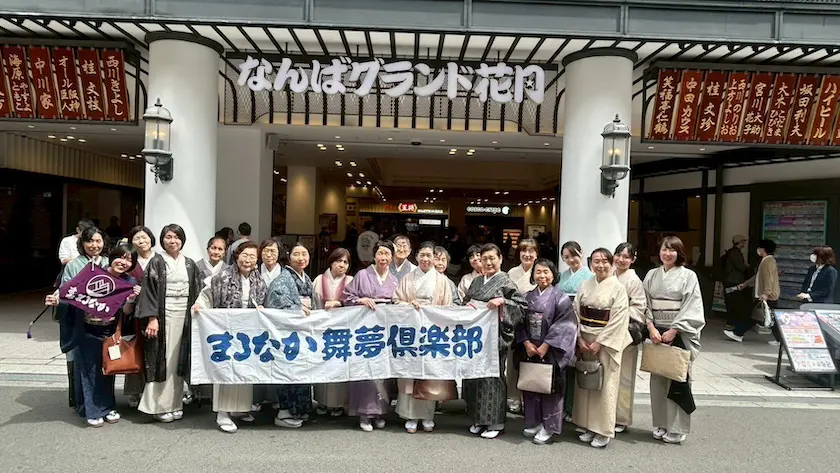 日本の伝統的な着物を着た一団が、日本語の看板が掲げられた劇場の入り口の前に笑顔で立っている。彼らは日本語の文字が書かれた大きな横断幕と旗を持ち、「楽しむ会」を祝っている。お祭り気分はスタッフBLOGにぴったりだ。.