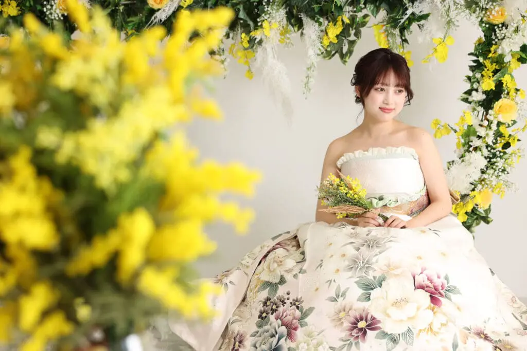 花嫁衣装を思わせる花柄のオフショルダードレスを着た女性が、黄色い小さな花束を抱えて座っている。緑豊かなアーチに囲まれ、鮮やかな花が咲き乱れる光景は、今月のスタッフBLOGにぴったりだ。.