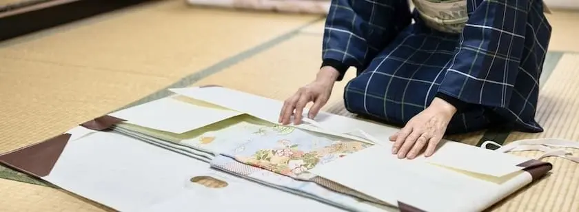青い格子柄の着物を着た人が、畳の上で花柄のパステルカラーの着物を丁寧にたたみ、スタッフBLOG用に白いティッシュペーパーで包む。.