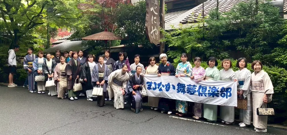 京都の路上で、日本の伝統的な着物を身にまとった女性たちが、日本語の文字が書かれた白と青の横断幕を掲げてポーズをとっている。緑の木々や瓦屋根の建物が、夏の風物詩ならではの穏やかで祝祭的な魅力を醸し出している。.
