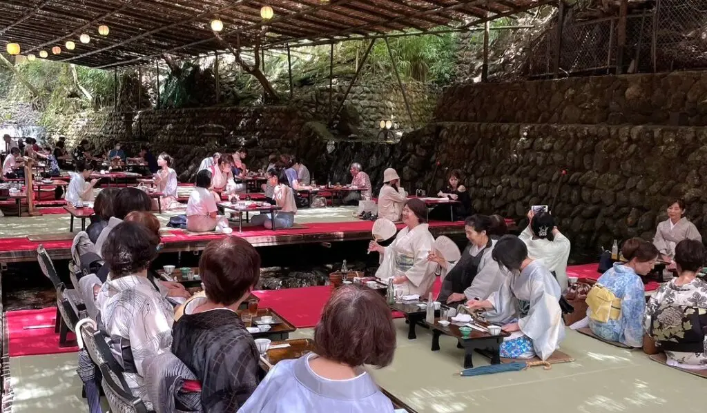 和服姿の人々が、低いテーブルを囲んで畳の上に座り、竹の屋根の下で屋外での食事を楽しんでいる――その光景は、緑豊かな木々や石垣が、夏の風物詩とも言える穏やかな雰囲気を醸し出す「京都の川床」を彷彿とさせる。.
