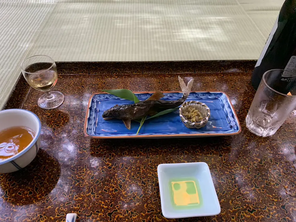青い長方形の皿に、付け合わせを添えた焼き魚が盛り付けられ、夏の風物詩の魅力を存分に引き立てている。その傍らには、漬物、お茶、白ワイン、緑色のタレなどが置かれており、すべてが、背景に敷物が敷かれた模様入りのテーブルの上に、芸術的に盛り付けられている。.