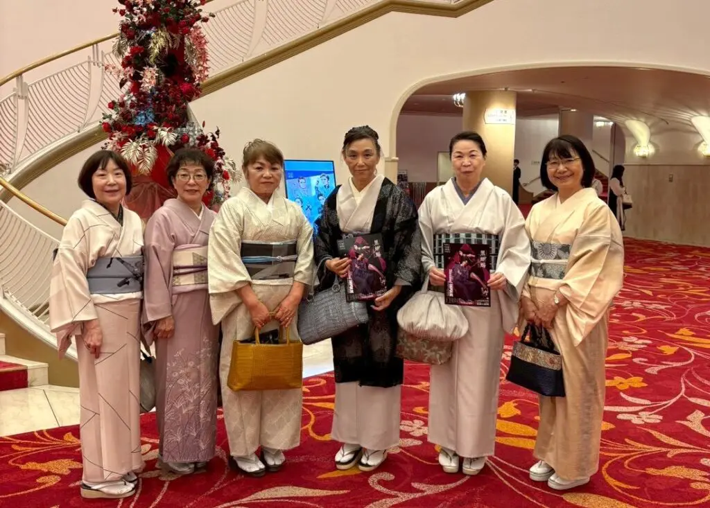 白い壁、螺旋階段、そして背の高いフラワーアレンジメントが配された豪華なロビーで、上品なパステルカラーや柄物の着物をまとった6人の女性が、赤いカーペットの上に並んで立っている。このスタッフブログでは、そのうちの2人がバレエダンサーが表紙を飾ったプログラムを手にしており、全員がカメラに向かって笑顔を見せている。.