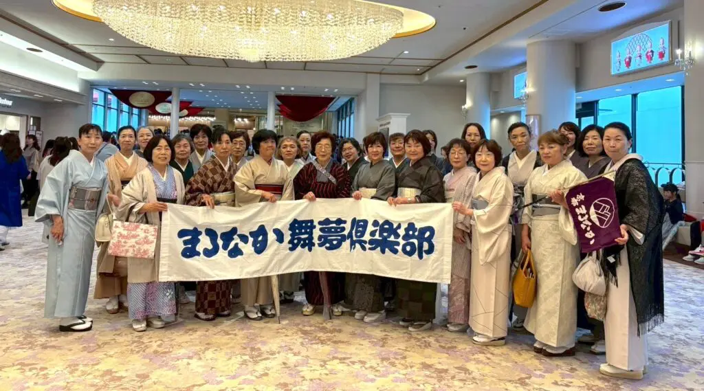 伝統的な着物を着た女性たちが屋内に立ち、青い日本語の文字が書かれた白い横断幕を手に、笑顔で写っている。大きな窓と花柄のカーペットが敷かれた優雅な部屋のシャンデリアの下で、彼女たちはスタッフブログで紹介される「お出掛け」の準備万端のようだ。.