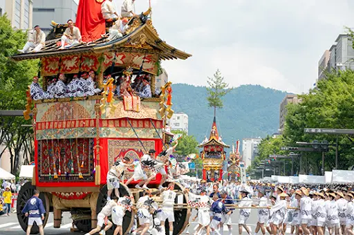 活気あふれるパレードは、まさに「夏の風物詩」そのもので、赤と金で彩られた華やかな木製の山車が、日本の伝統衣装を身にまとった人々を乗せて、群衆に引かれながら街中を練り歩きます。木々や山々が、このお祭りの光景を美しく彩っています。.