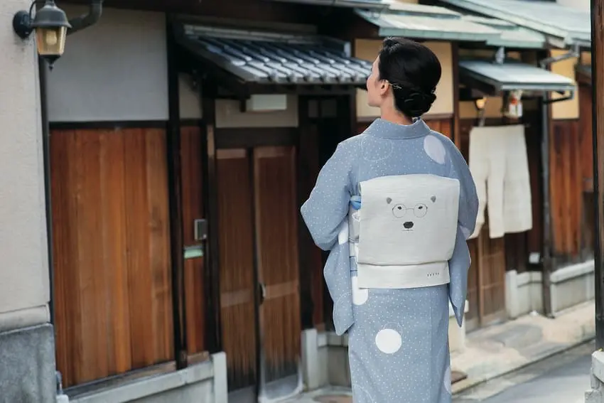 白い水玉模様の青い着物を着た女性が、木造の建物が立ち並ぶ通りを歩いている。 彼女は黒髪を髷にまとめ、淡い色の帯を締めている。その帯にはクマの顔がシンプルに描かれており、このチャーミングなアクセントが『夏の小物』の遊び心あふれる雰囲気を彷彿とさせる。この光景からは、当スタッフのブログによく綴られる穏やかなひとときのように、静けさと伝統の雰囲気が漂っている。.