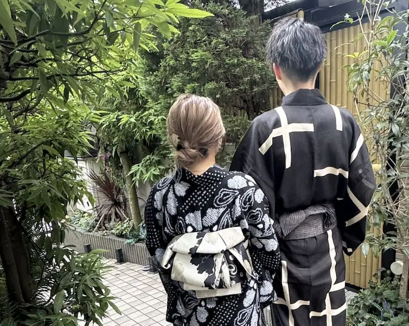 屋外で2人が並んで立っている後ろ姿が写っています。2人は日本の伝統的な浴衣を着ており、女性は花柄の浴衣に装飾的な帯を、男性は大胆な白い線柄の浴衣を身につけています。周囲は青々とした緑と竹の垣根に囲まれており、当サイトのスタッフブログで紹介されている「お出かけ」にぴったりの、穏やかな雰囲気を醸し出しています。.