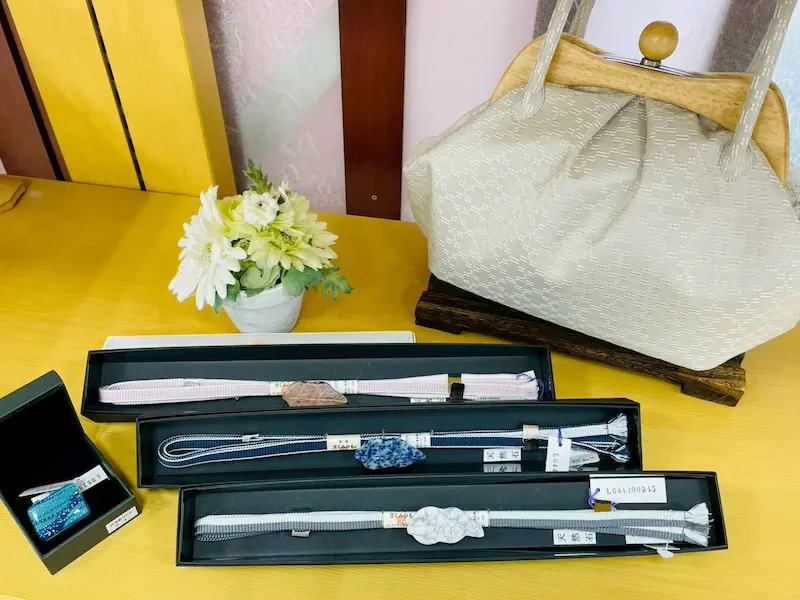 木製のテーブルの上には、ベージュのテクスチャーが施されたハンドバッグが、小さな白い花束の隣に直立して置かれています。お出かけにぴったりですし、浴衣のコーディネートを引き立てるのにも最適です。バッグの前には、当店のスタッフブログでも紹介されている通り、黒い箱に収められた3本の装飾用着物帯の帯締めと、ターコイズ色のアクセサリーが並んでいます。.