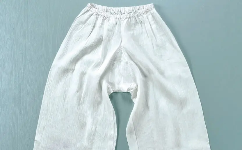 ゆったりとしたシルエットの白いコットンまたはリネンの股割れパンツが、水色の背景の上に平らに並べられています。このパンツはウエストにゴムが入っており、裾が広めのデザインで、股の部分に開いた縫い目があります。夏の必須アイテムとして、涼しく過ごすのに最適です。その他のスタイリングのヒントや撮影の裏側については、スタッフブログをご覧ください。.
