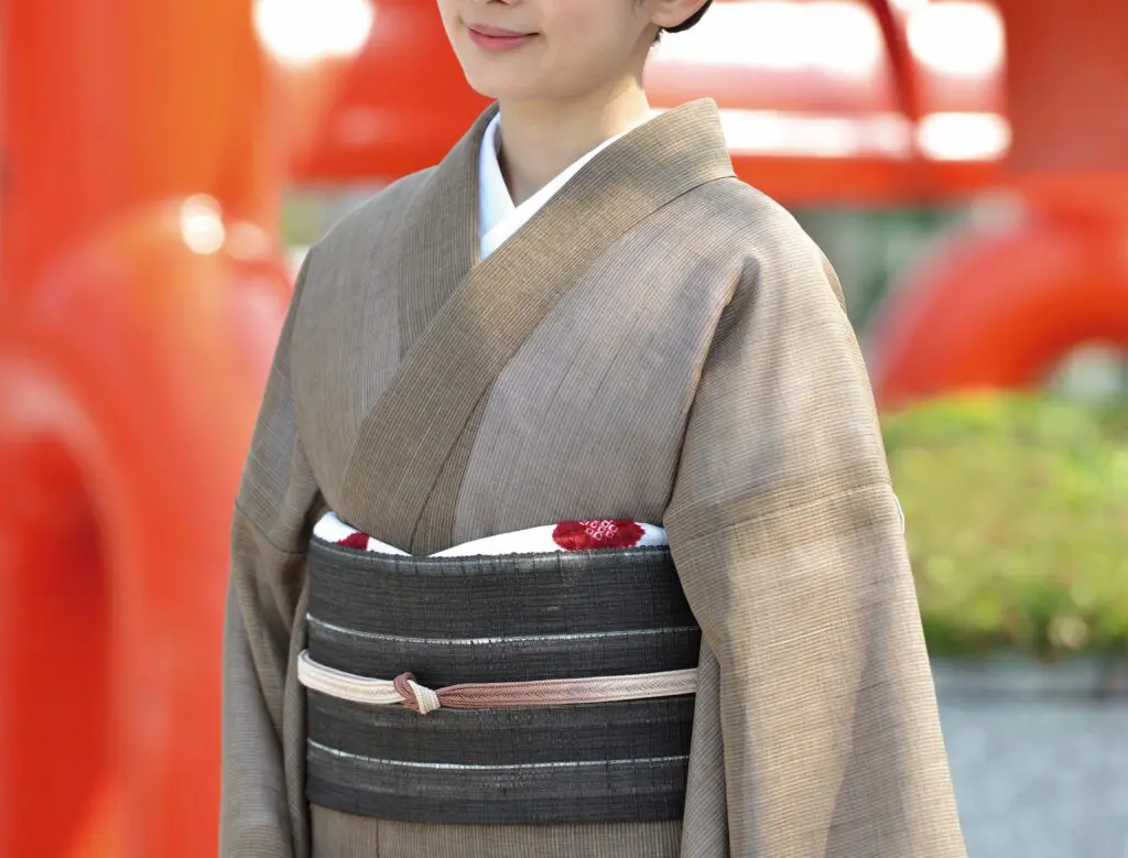 控えめな縞模様の入った茶色の着物――おそらく「全国紬」で作られたもの――と、暗い色の柄入りの帯を身に着けた人物。顔の下半分と上半身のみが見えている。背景は赤みがかったオレンジ色の建造物や緑がぼやけて描かれており、スタッフBLOGの記事によく登場する雰囲気を彷彿とさせる。.
