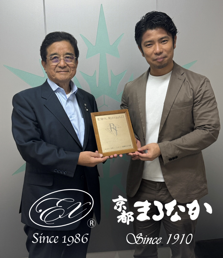 2人の男性が並んで立ち、笑顔で一緒にプラークを掲げています。1人はメガネをかけ、ダークスーツに青いシャツを着ており、もう1人は白いシャツの上にベージュのスーツを着ています。 二人の背後には、下部に日本語と英語のテキストが記載されたスタイリッシュなロゴがあり、この場面が「ジュエリー相談会」に関連しているか、あるいは「スタッフブログ」で取り上げられていることを示唆している。.