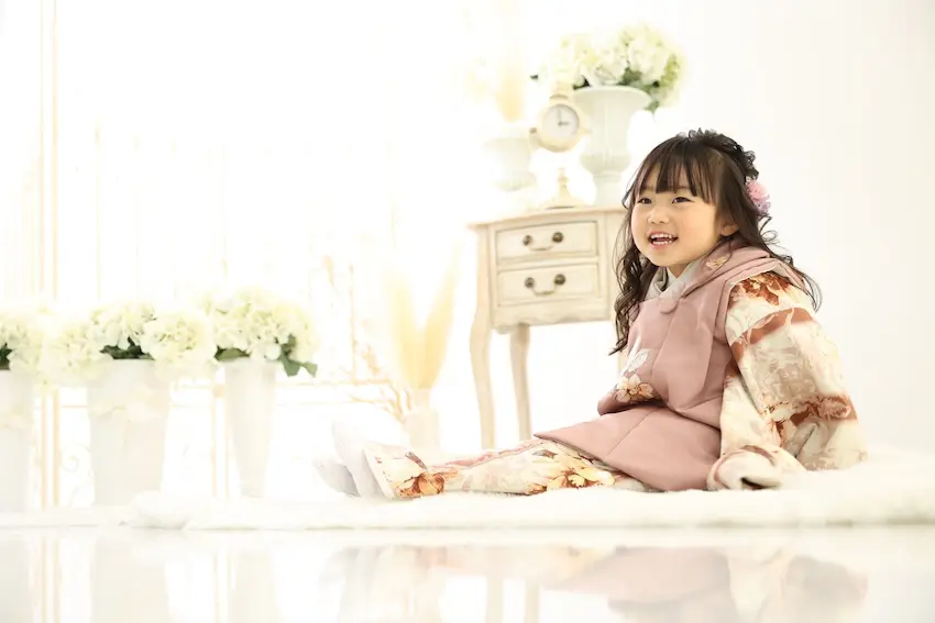 花飾りを髪に挿した黒髪の少女が、淡いピンクの柄物着物を着て微笑んでいる。彼女は、白い花のアレンジメントが飾られ、背景にはヴィンテージ風の小さな木製テーブルが置かれた、明るくシンプルな部屋の中で、白いラグの上に座っている。.