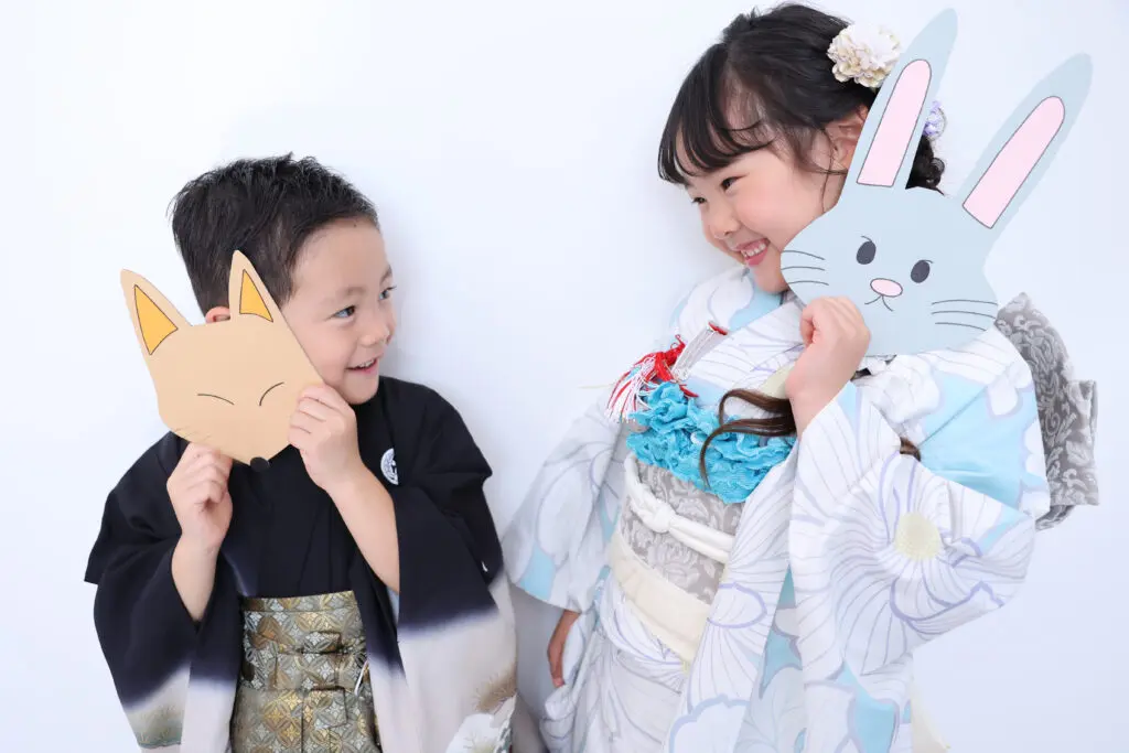 白い背景を背に、日本の伝統的な着物を着た少年と少女が互いに微笑み合っている。少年は顔に狐の仮面をかざし、少女はウサギの仮面を手に持っている。二人の子供は、お祝いの席にふさわしい、柄入りの正装を身にまとっている。.