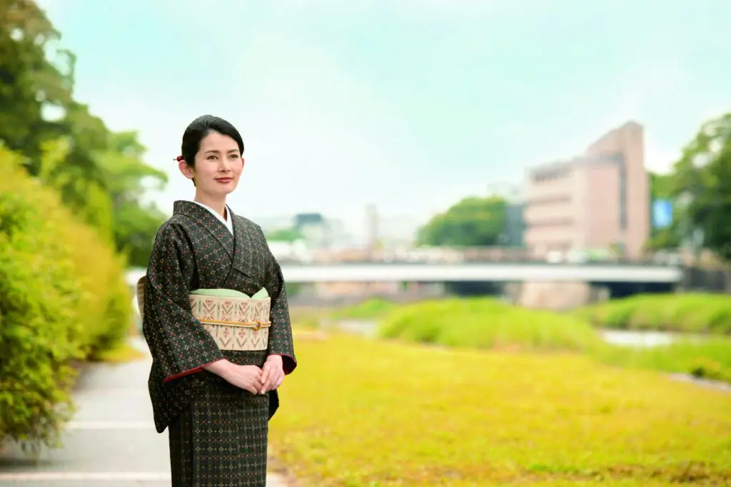 秋の装いをした女性が、草や低木に囲まれた小道に立っており、背景には川と橋が見えます。青空の下には木々や大きな建物が見えます。彼女は両手を胸の前で組んで、ほほえんでいます。#スタッフBLOG.