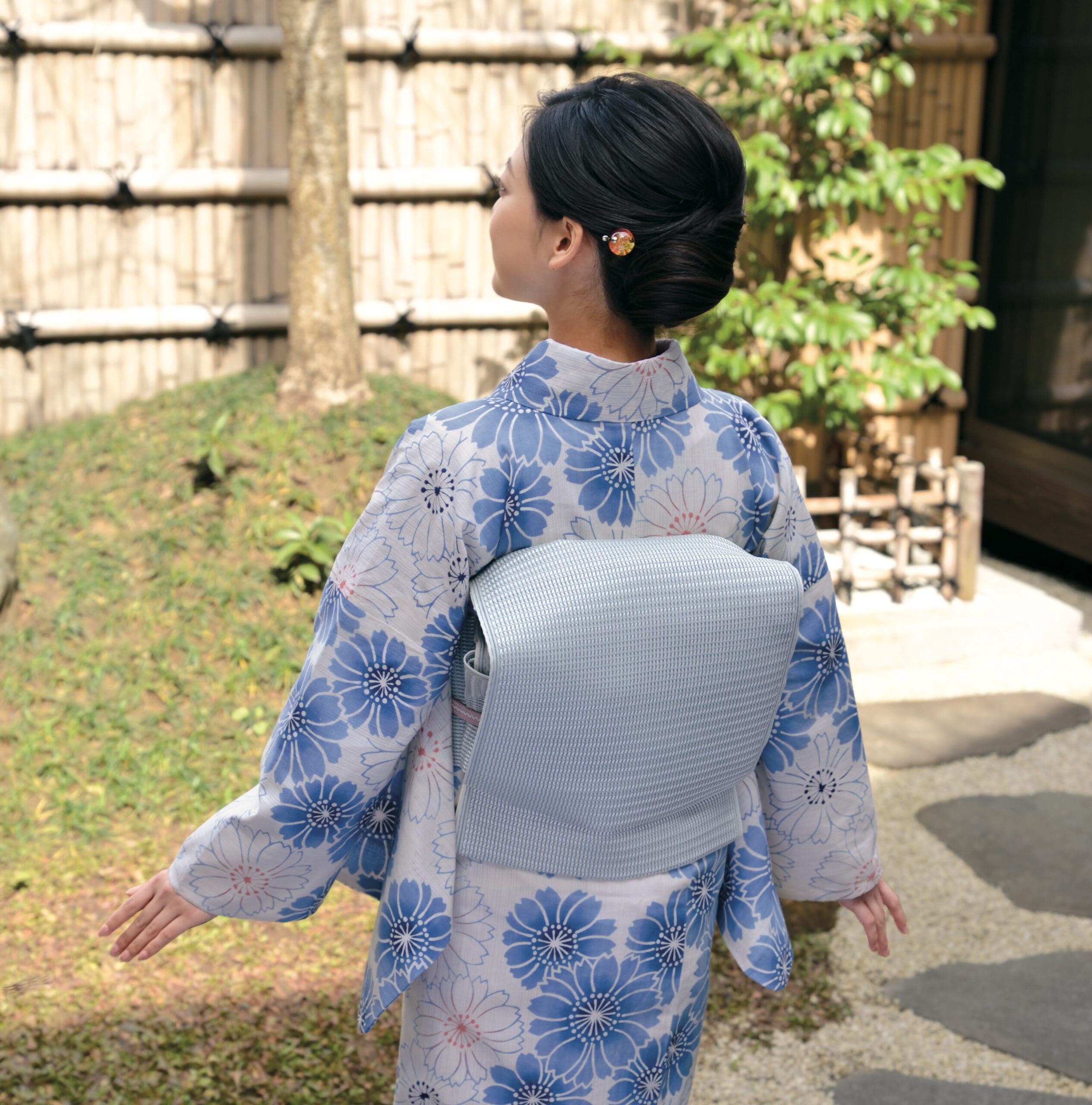 大きな花柄があしらわれた水色の着物を着た人が屋外に立っており、カメラから背を向けています。髪は小さなヘアピンできちんとセットされています。竹垣、木、緑の茂みがある静かな環境です。この美しい瞬間は、スタッフBLOGで紹介されている着物クリニックキャンペーンの一部になるかもしれません。