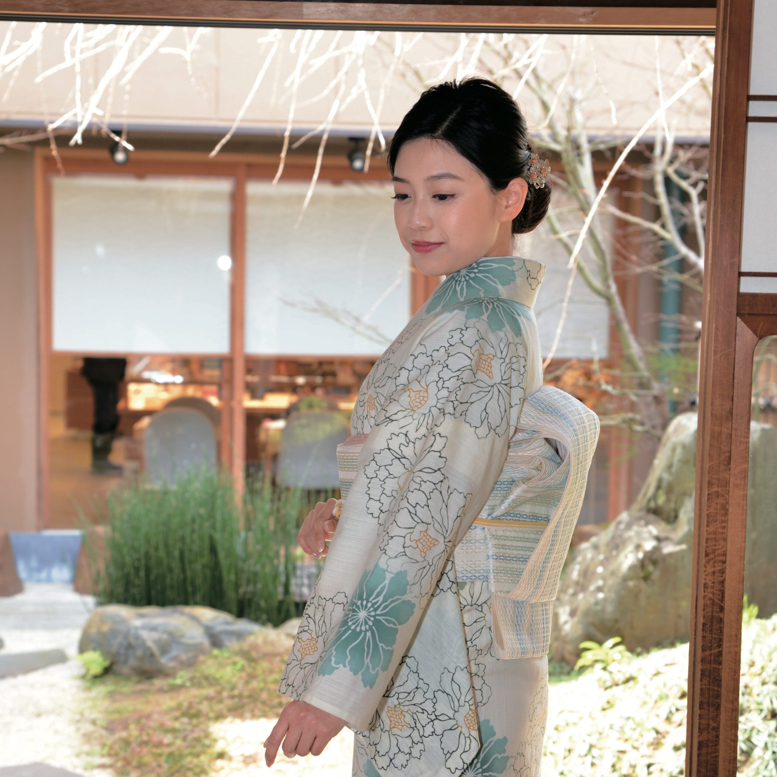 浴衣に似た花柄の伝統的な着物を着た女性が、室内の窓際に立っています。彼女の髪はきちんとセットされ、ヘアピンで飾られています。窓越しに、背景に岩と緑のある庭園が見えます。