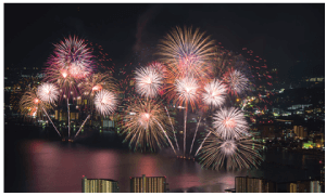 鮮やかな花火が打ち上がる水辺の街の夜景。赤、白、金など色とりどりの花火が爆発し、暗い空を照らし、水面に映ります。背景の街並みは、建物や通りの明かりで照らされています。