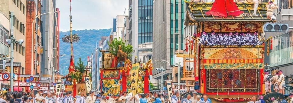 街路を練り歩く賑やかな日本の伝統的な祭りパレードでは、精巧なデザインで飾られた鮮やかな木製の山車が登場します。夏の装いの参加者が山車を曳き、近代的なビル群と遠くの山々が対照的な背景を作り出します。詳細はスタッフBLOGをご覧ください。