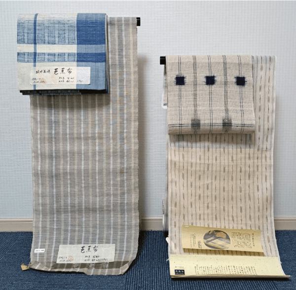 白い壁に縦に巻かれた 2 つの布地。左のロールには灰色の縞模様と文字の入ったラベルが付いており、その上に青と白の折り畳まれた布地があります。右のロールはベージュのベースに縦線と青い四角い模様があり、下部近くに文字と画像の部分があります。