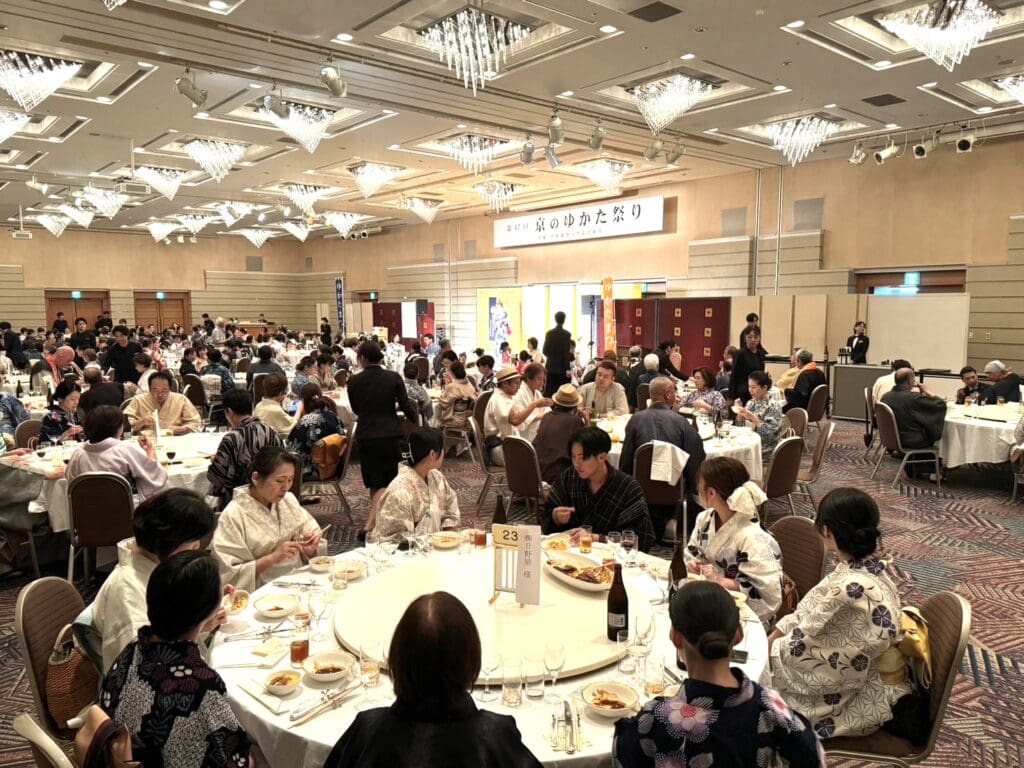 大きな宴会場には丸テーブルが並び、それぞれに複数のゲストが着席して食事を楽しんでいます。出席者のほとんどは伝統的な日本服を着ています。天井からはエレガントなシャンデリアが吊り下げられ、正面にはバナーが掲げられたステージがあります。雰囲気は活気があり、社交的です。