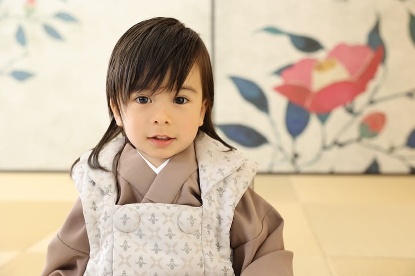 長い黒髪の子供が七五三のお祝いに着る和服を着ています。子供は花柄の飾り壁を背景に畳の上に座り、穏やかな表情でカメラを見ています。スタッフブログで詳しくご覧ください！