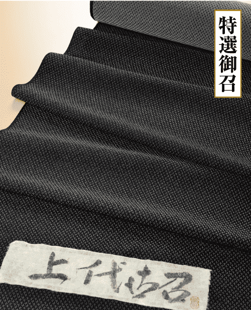 繊細なテクスチャ模様の黒い布を折りたたんだ状態のクローズアップ。右上隅の白いラベルに縦書きの漢字が 2 つ印刷され、左下隅のベージュ色のラベルにも漢字が書かれています。布地は滑らかで上品な外観です。.