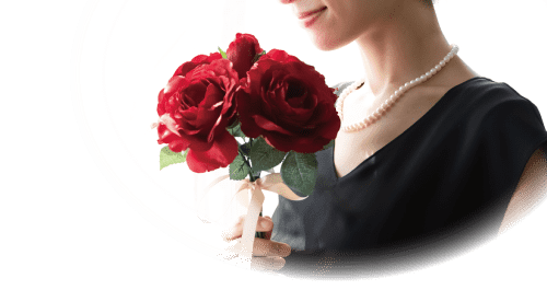 黒いドレスを着て真珠のネックレスをした女性が、緑の葉が付いた鮮やかな赤いバラの花束を持っています。背景は柔らかく照らされており、穏やかで穏やかな雰囲気が醸し出されています。画像は部分的に円形に切り取られており、花束と女性の穏やかな表情に焦点が当てられています。.