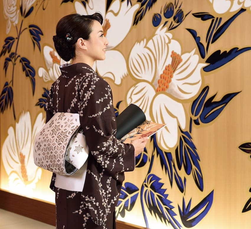 茶色の花柄の着物と白い帯を締めた人物が、大きな花の壁画に向かって黒いノートとパンフレットを持っています。この壁画は、木の背景に濃い青の葉が付いた白い蘭が描かれており、スタッフブログの感動的なエントリーにぴったりの、エレガントで穏やかな雰囲気を醸し出しています。.