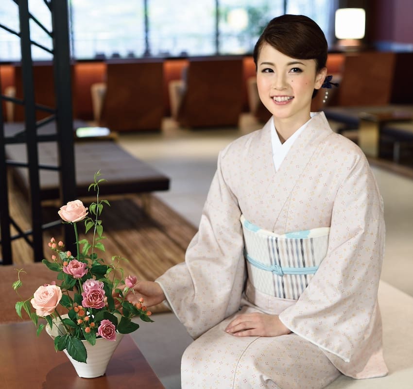 淡い柄の着物を着た女性が店内に座り、微笑んでいる。淡いブルーと白の帯を上手に結び、涼しい季節に備えて落ち着いた優雅さを漂わせている。彼女の横には、ピンクのバラと緑の花瓶が置かれ、大きな窓に囲まれたモダンな座席エリアに魅力を添えている。#スタッフBLOG.