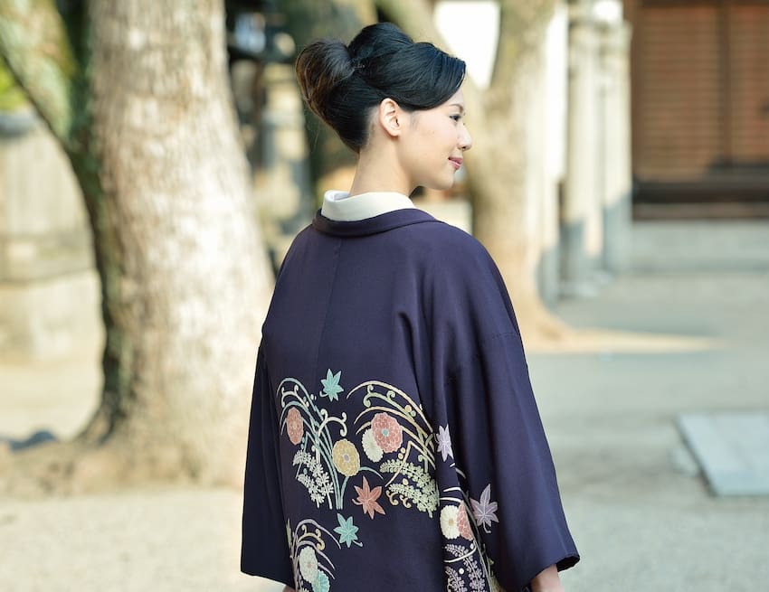 複雑な花と葉の模様が描かれた紫色の着物を着た女性が屋外に立って横を向いています。彼女の髪は上品なアップスタイルになっています。背景にはぼやけた木の幹と建物の壁があり、この穏やかな日本の風景の中で彼女が防寒の準備をしている様子が捉えられています。.