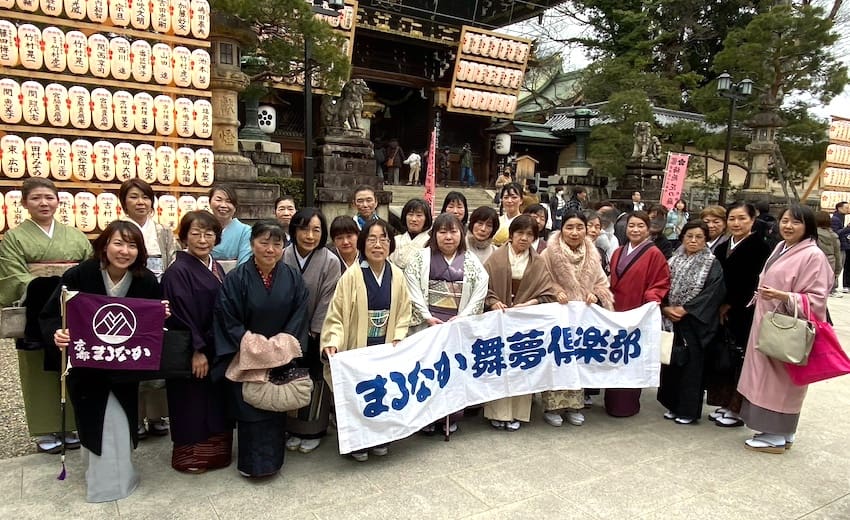 日本の伝統的な衣装を着た大勢の人々(ほとんどが女性)が屋外に立っています。彼らは日本語の文字が書かれた 2 つの旗を持っています。彼らの後ろには、提灯の列で飾られた装飾的な構造物があります。背景には木々と追加の構造物が見えます。.