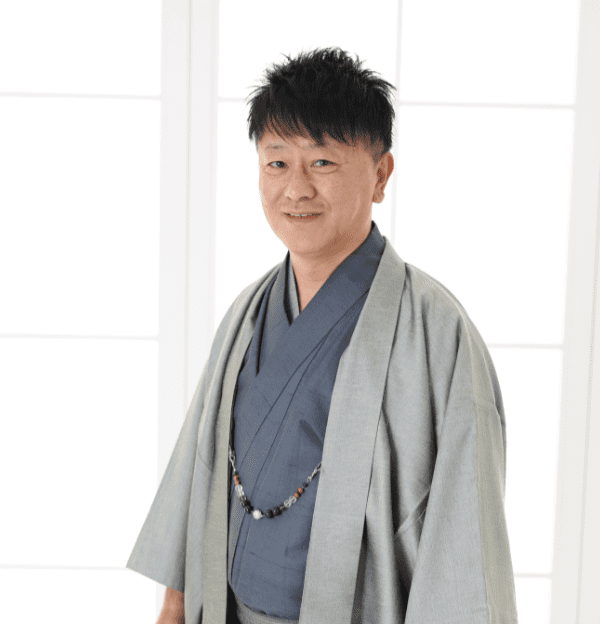 日本の伝統的な着物を着た人物が、白い障子風の背景の前に立っています。着物はグレーで、内側は濃い色で、ビーズのネックレスをアクセサリーとしてつけています。人物は短くて黒い髪をしており、カメラに向かって優しく微笑んでいます。.