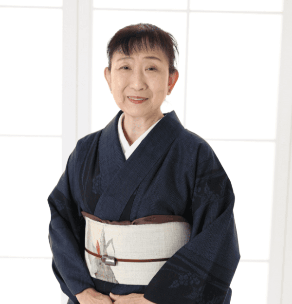 年配の女性が微笑んでいる。淡い色の細かな模様の着物に、小さな模様の入った淡い色の帯を締めている。髪はきちんと後ろに束ねられており、障子が張られた柔らかな光が差し込む窓の前に立っている。.
