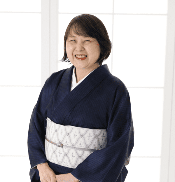 明るい窓の前に、淡い模様の帯を締めた濃紺の着物を着た女性が微笑みながら立っています。背景は柔らかく照らされ、伝統的な衣装が際立っています。彼女は短い黒髪で、両手を優しく前で組んでいて、楽しそうに見えます。.