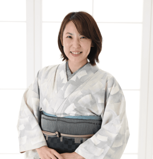 明るい逆光に照らされた窓の前に立って微笑む人物。淡い色の繊細な模様の着物と、濃い色の市松模様の帯を締めている。背景は柔らかく照らされ、人物の周りに穏やかな輝きを放っている。.