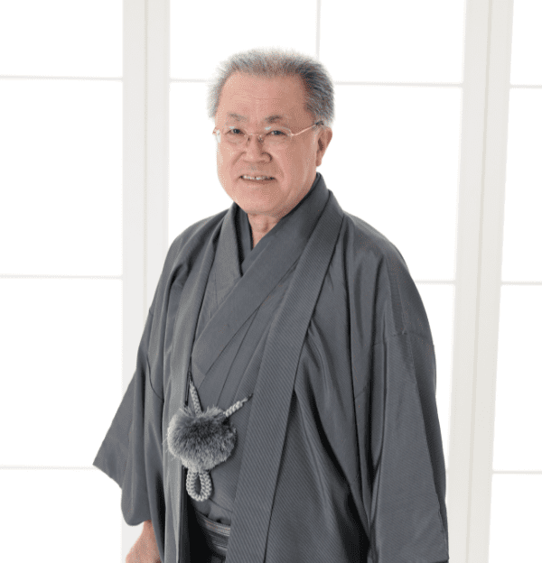 腰に飾り紐を巻いた伝統的な灰色の着物を着た年配の男性が微笑みながら立っている。彼は灰色の短い髪と眼鏡をかけている。背景には白い格子状のパネルを通して明るく拡散した光が映っている。.