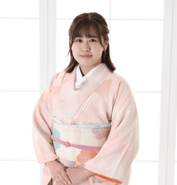 淡いピンク色の花柄の着物と華やかな帯を身に着けた人物が、室内の窓の前に立っています。背景は、格子模様の白い窓から差し込む柔らかな光で明るくなっています。.