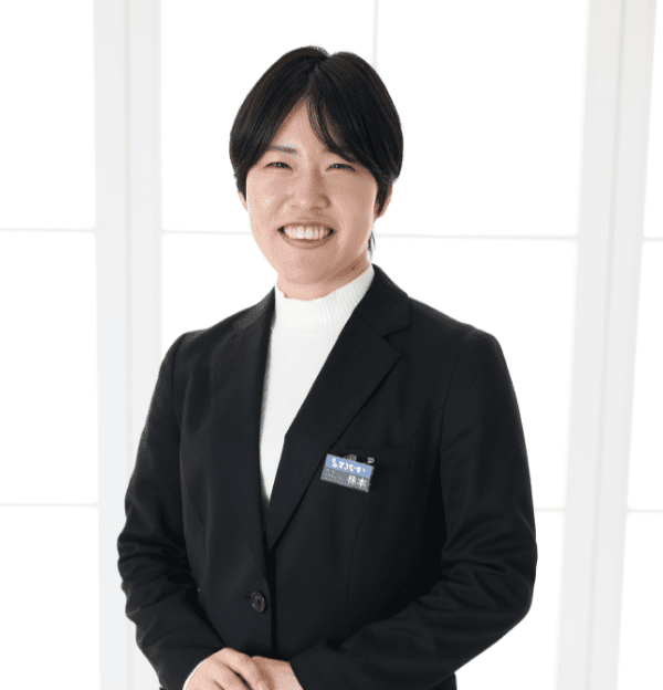 黒いブレザーと白いタートルネックを着て、笑っている短い黒髪の人物。ブレザーには名札が付いています。背景は白いパネルで柔らかく照らされています。.