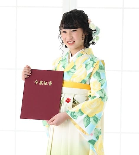 花柄の着物を着た若者が、金色の日本語の文字が書かれた栗色の認定証のフォルダーを持っています。着物には黄色と緑の花が描かれ、帯は黄色です。彼らは明るく柔らかな光を放つ背景を背に微笑んでいます。.